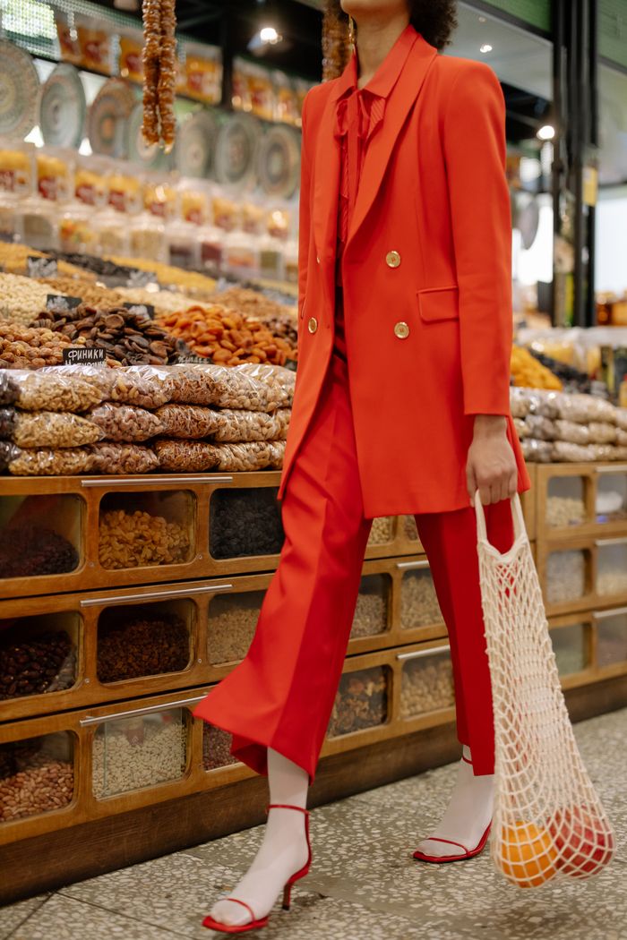 A fashionable woman in a red blazer shops for nuts in a market carrying a mesh bag.